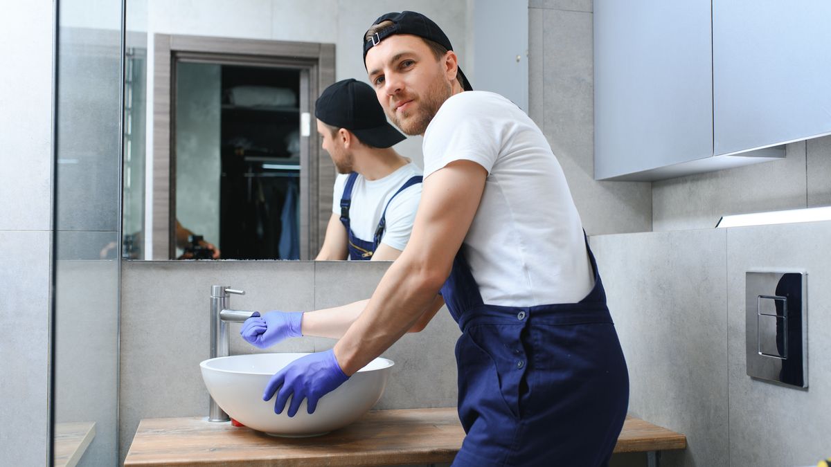 Plumber in overalls and gloves fitting a basin in a grey tiled bathroom
