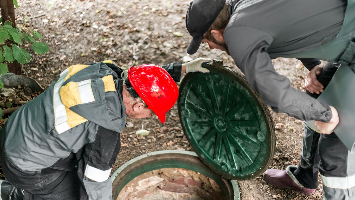 Two workers in overalls looking down into an open manhole to check the sewer line